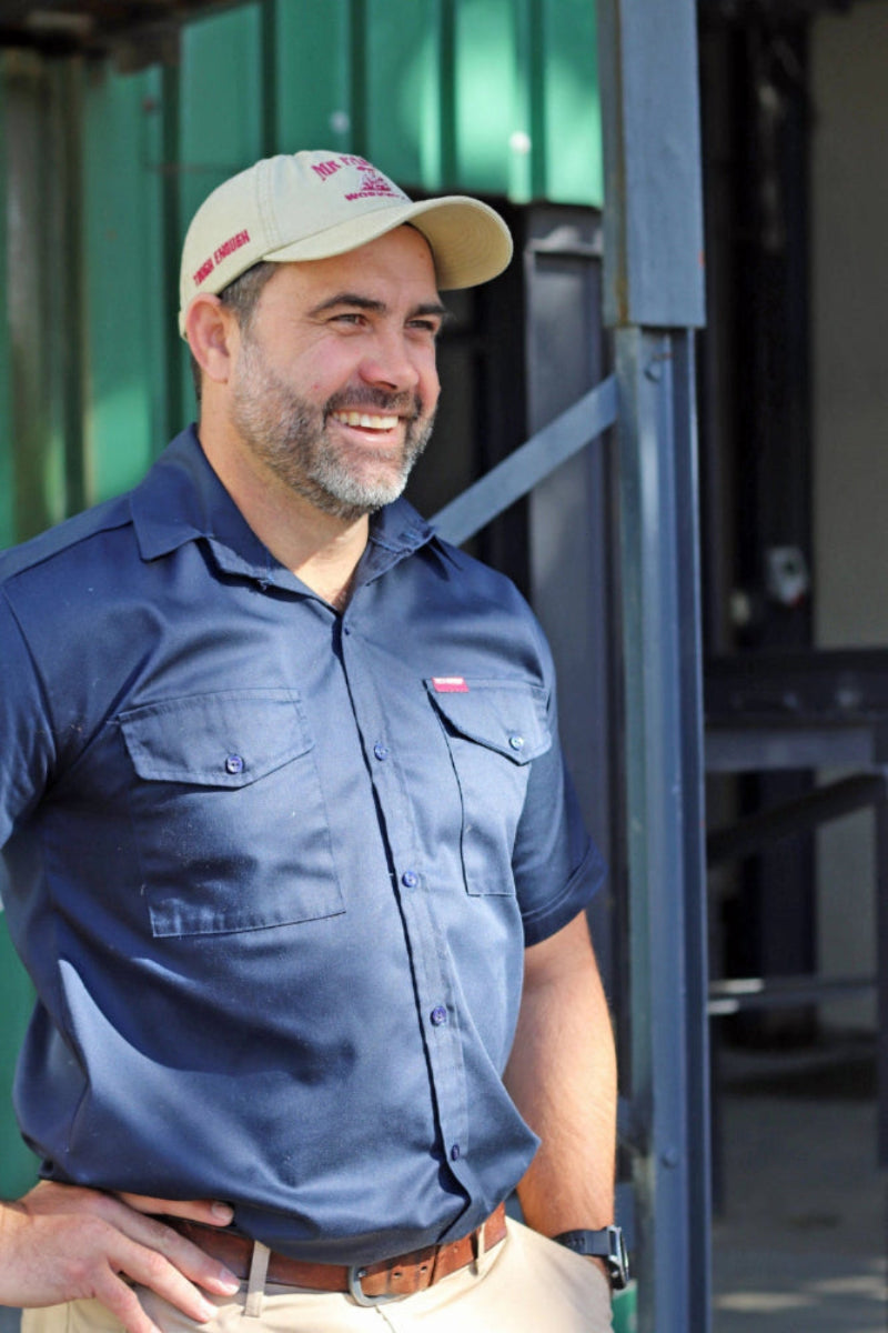 A smiling man wearing a navy blue short-sleeve button-up shirt and a beige Mr Farmer Workwear cap, standing in front of a green corrugated metal wall and dark metal structure.