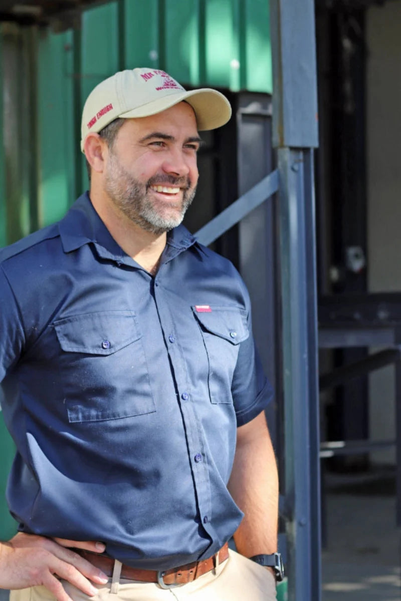 A smiling man wearing a navy blue short-sleeve button-up shirt and a beige Mr Farmer Workwear cap, standing in front of a green corrugated metal wall and dark metal structure.