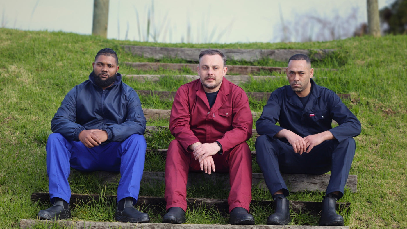 Three men wearing Mr Farmer Overalls sitting on steps outdoors with a grassy area and building in the background