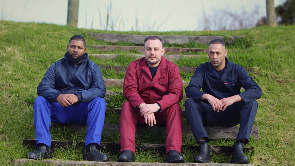 Three men wearing Mr Farmer Overalls sitting on steps outdoors with a grassy area and building in the background