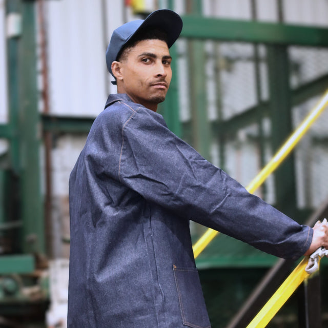 Person wearing a denim overall jacket and cap, holding a tool in an industrial setting