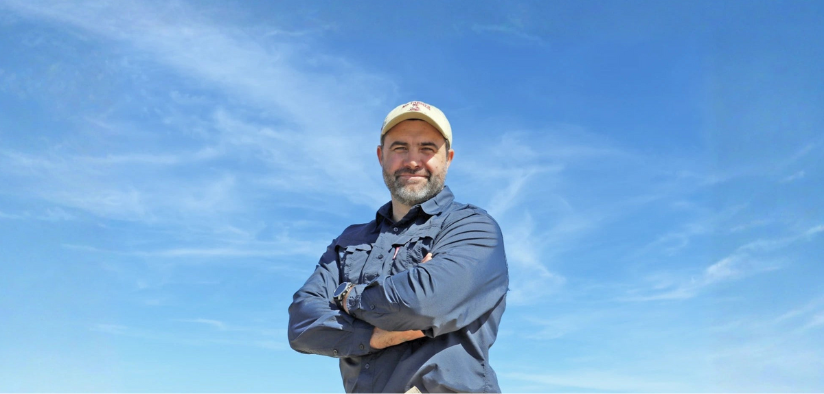 A man with a beard and a cap stands outdoors with his arms crossed, wearing a navy blue long-sleeve shirt and khaki pants against a bright blue sky with wispy clouds.