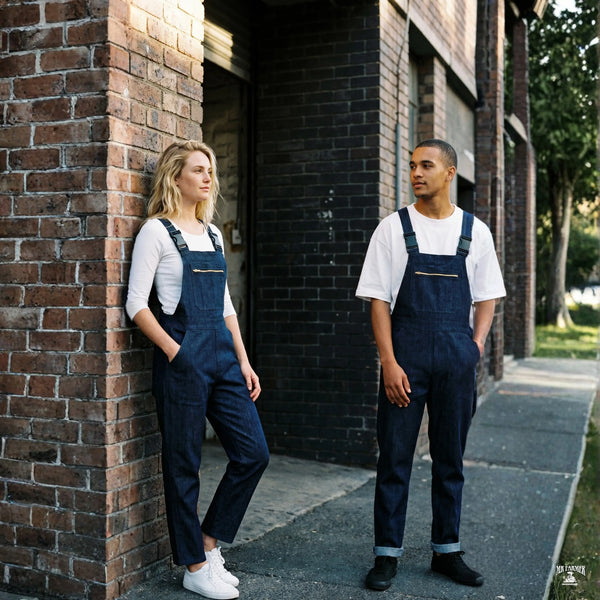 Two people wearing blue denim dungarees standing against a brick wall.