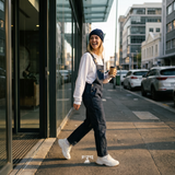 A woman walking on a city sidewalk wearing blue denim dungaree over a white long-sleeve shirt, a navy beanie, and white sneakers, holding a coffee cup.