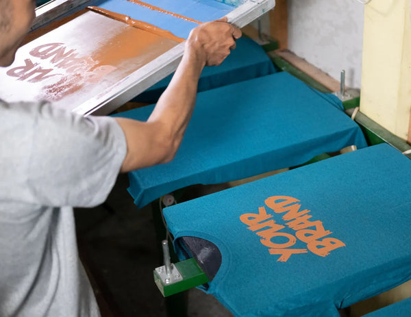 Person ironing blue t-shirts with visible branding in a workshop setting.